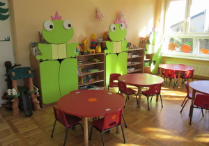 sala dla młodszych dzieci z zielonymi szafkami wykończonymi 3 żabkami oraz okrągłymi, czerwonymi stolikami